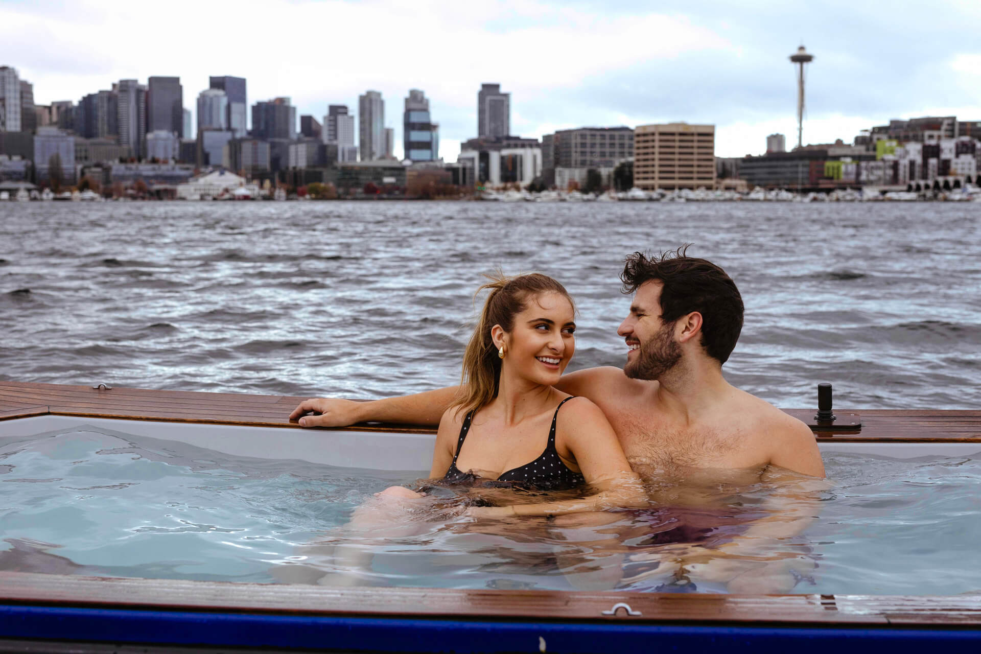 Photo of a man and woman sitting in a hot tub boat on a lake with the Seattle Space Needle in the background.