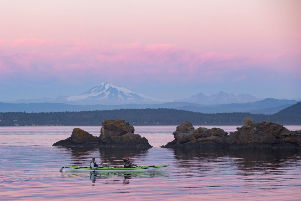 Two people in a double kayak enjoy a pink and purple sunset while kayaking in Bellingham Bay.