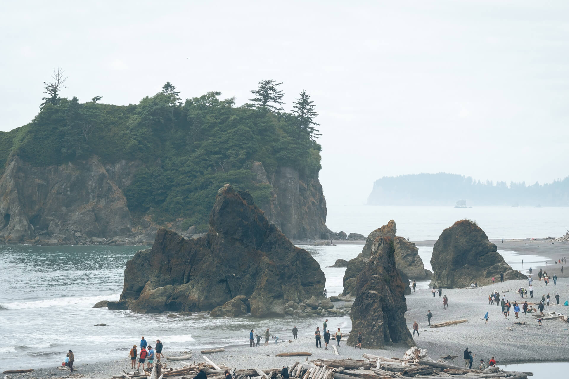 People walk along shore at Ruby Beach with jagged sea stacks in the background 