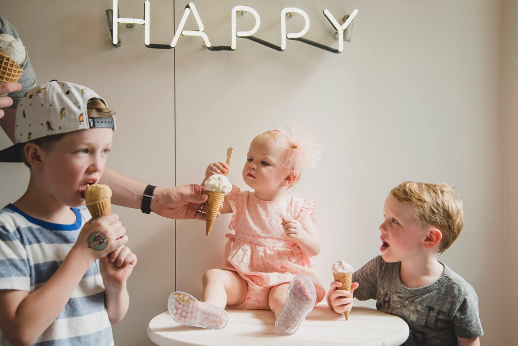 A toddler sits on a table eating an ice cream cone while two other young boys stand next to her eating ice cream cones