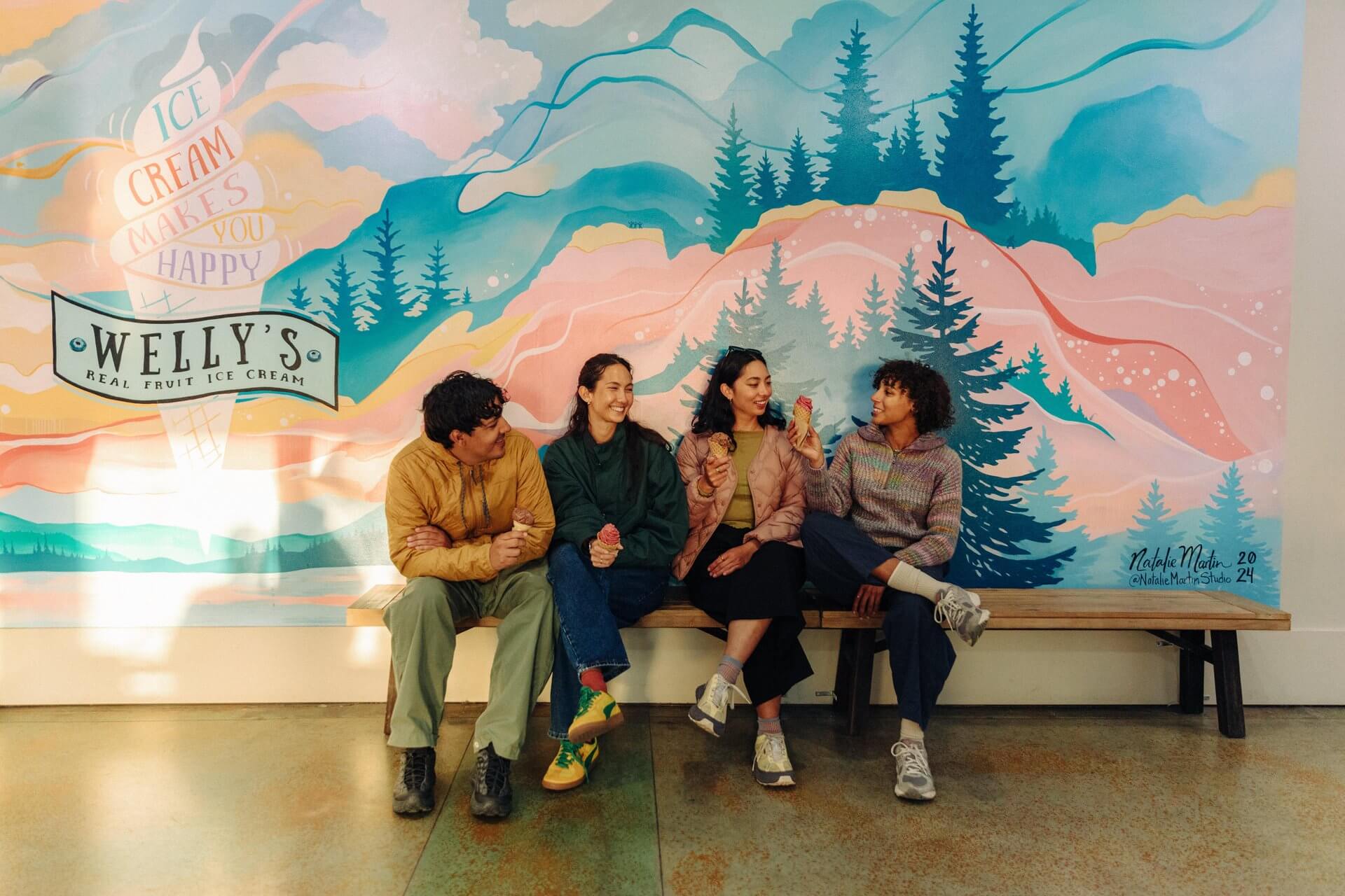 Four people sit on a bench in front of a mural and eat ice cream.