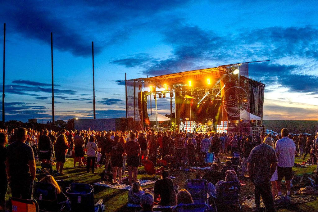 A photo of people standing and sitting on a lawn at sunset as they watch an outdoor concert