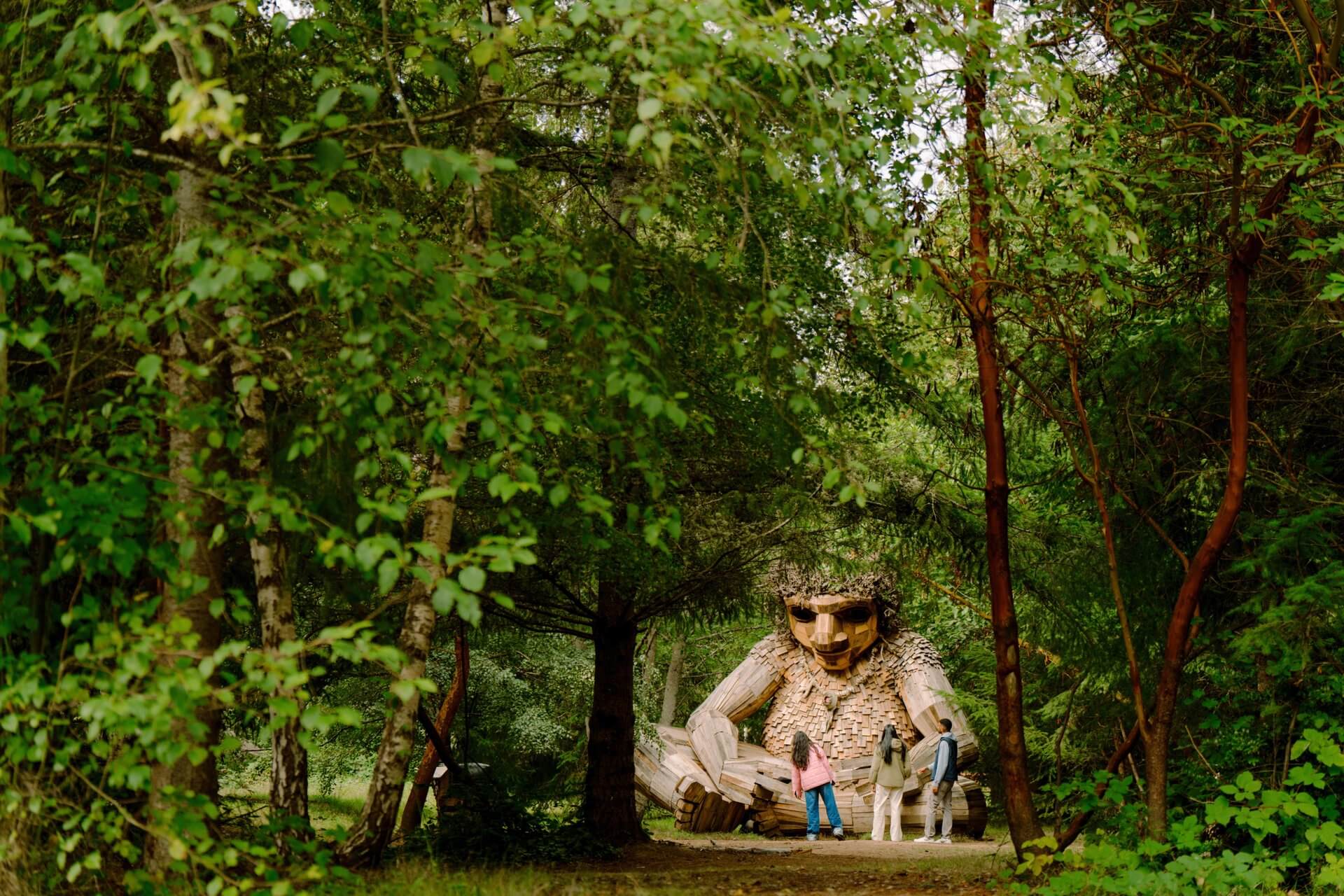Three people stand in front of a troll sculpture made of reclaimed wood.
